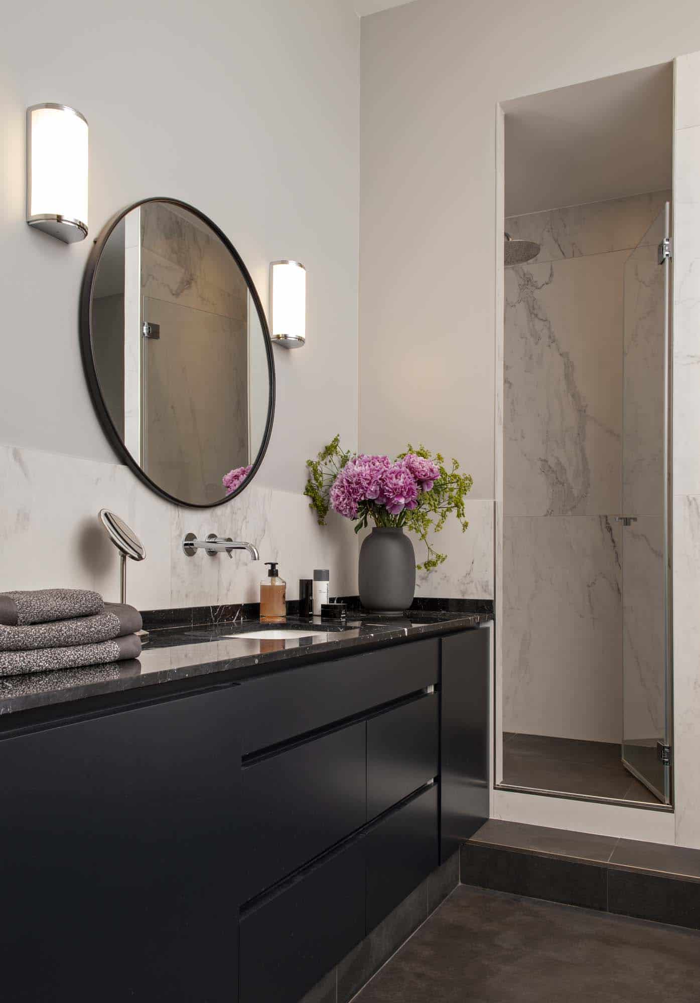 Salle de bains : meuble-vasque noir, miroir rond et douche à parois vitrées sur marbre clair
