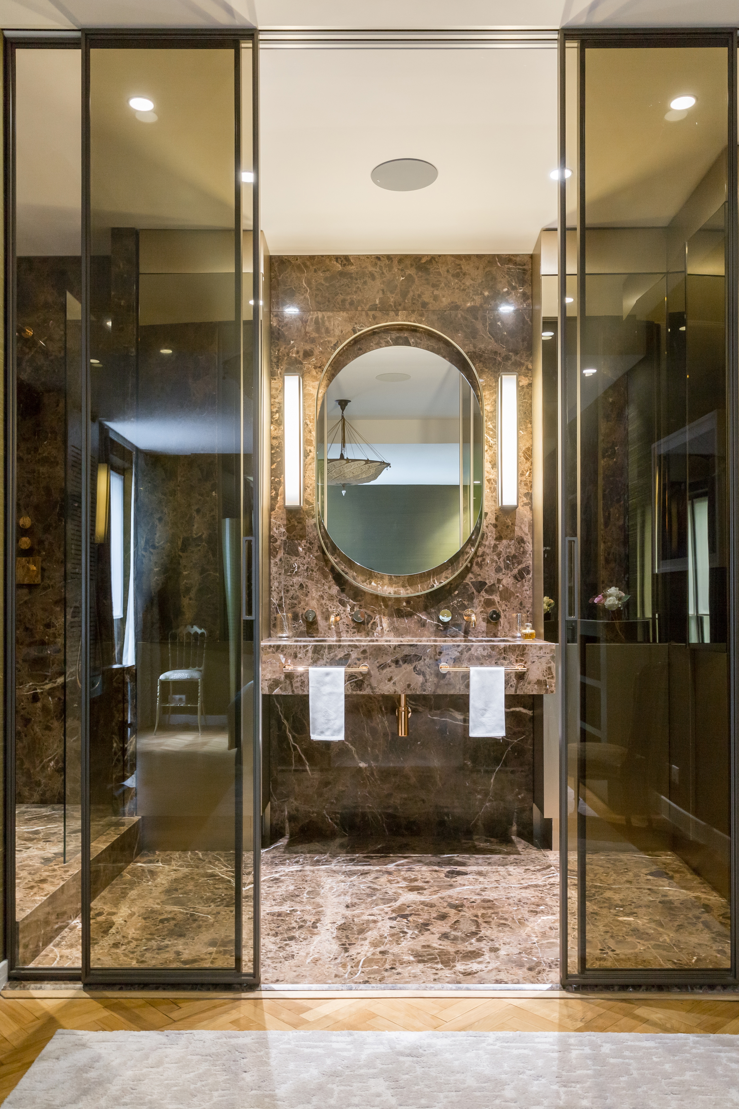 Salle d'eau en marbre derrière une paroi vitrée à cadre métallique, miroir ovale rétroéclairé