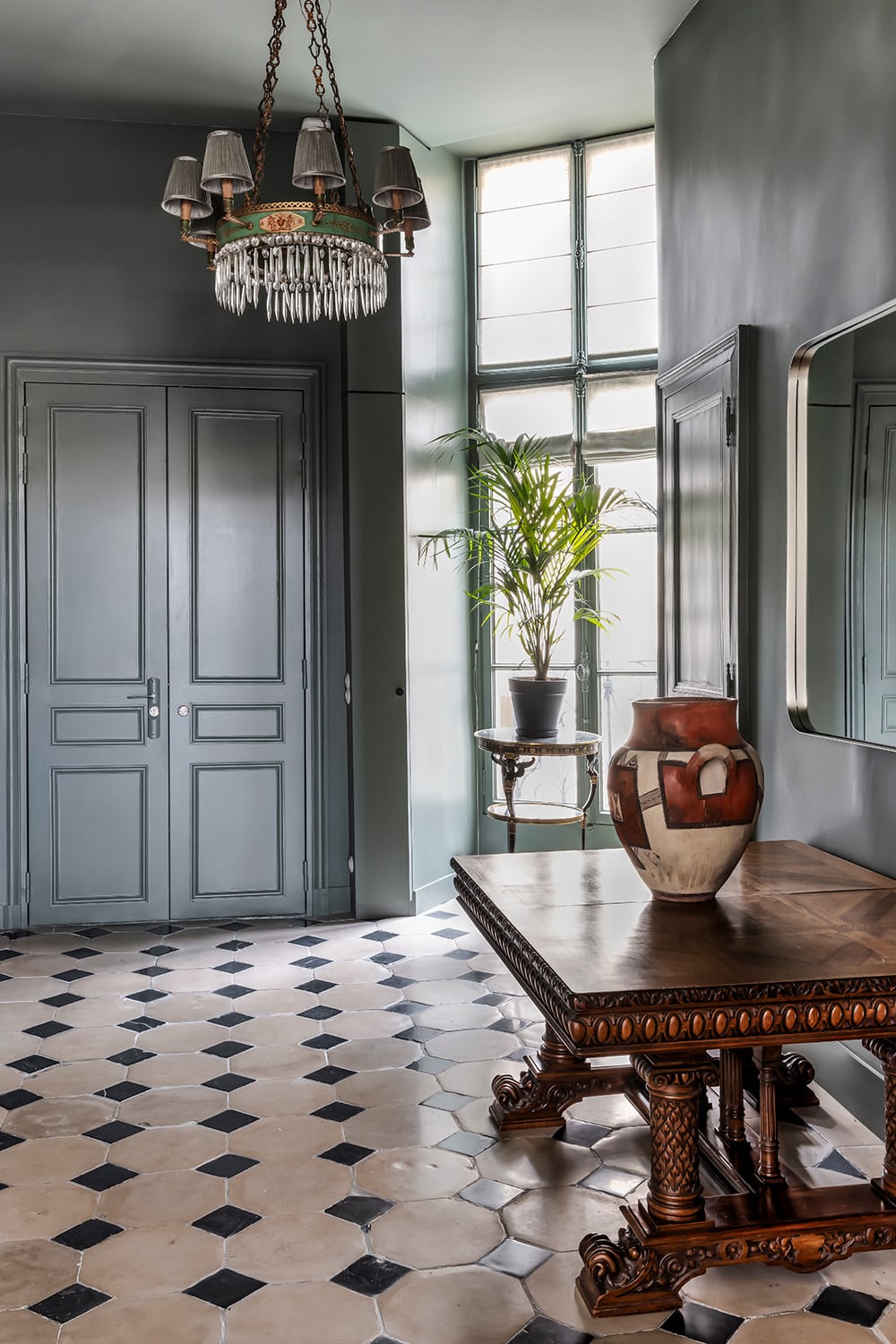 Entrée aux menuiseries gris-vert, portes doubles conservées et sol en damier sous une fenêtre haute
