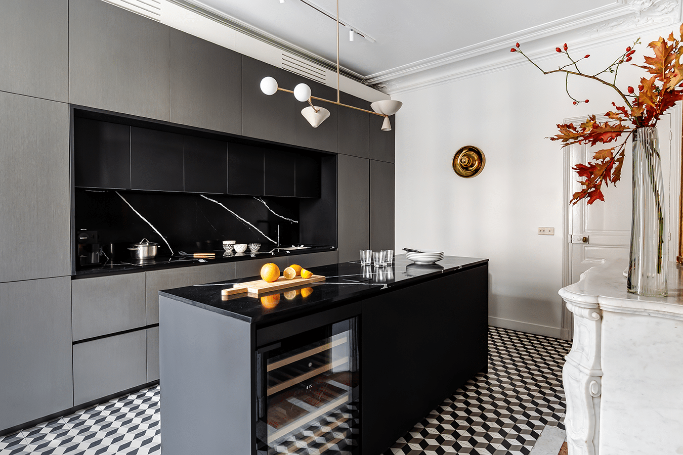 Grande cuisine noire : îlot central massif, façades sombres toute hauteur et sol en damier, ouverte sur l'enfilade de réception