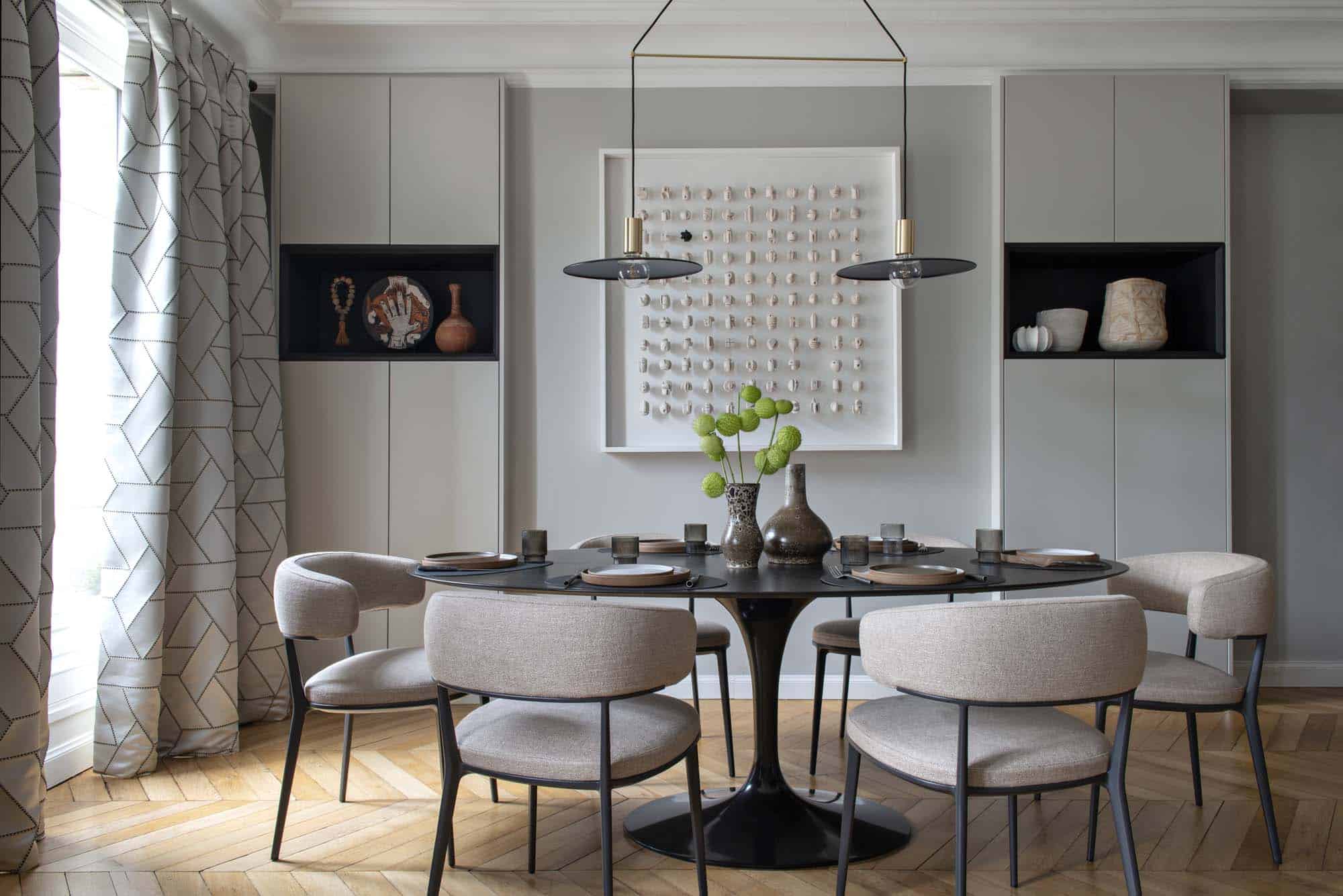 Salle à manger avec table ronde, niches sombres intégrées dans une paroi de menuiserie claire et double rideau